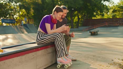 side view of a girl with a short haircut in a purple top striped pants in pink roller skates sits on a bench in a skate park and communicates with a brunette guy in a gray t-shirt with a phone in her hands