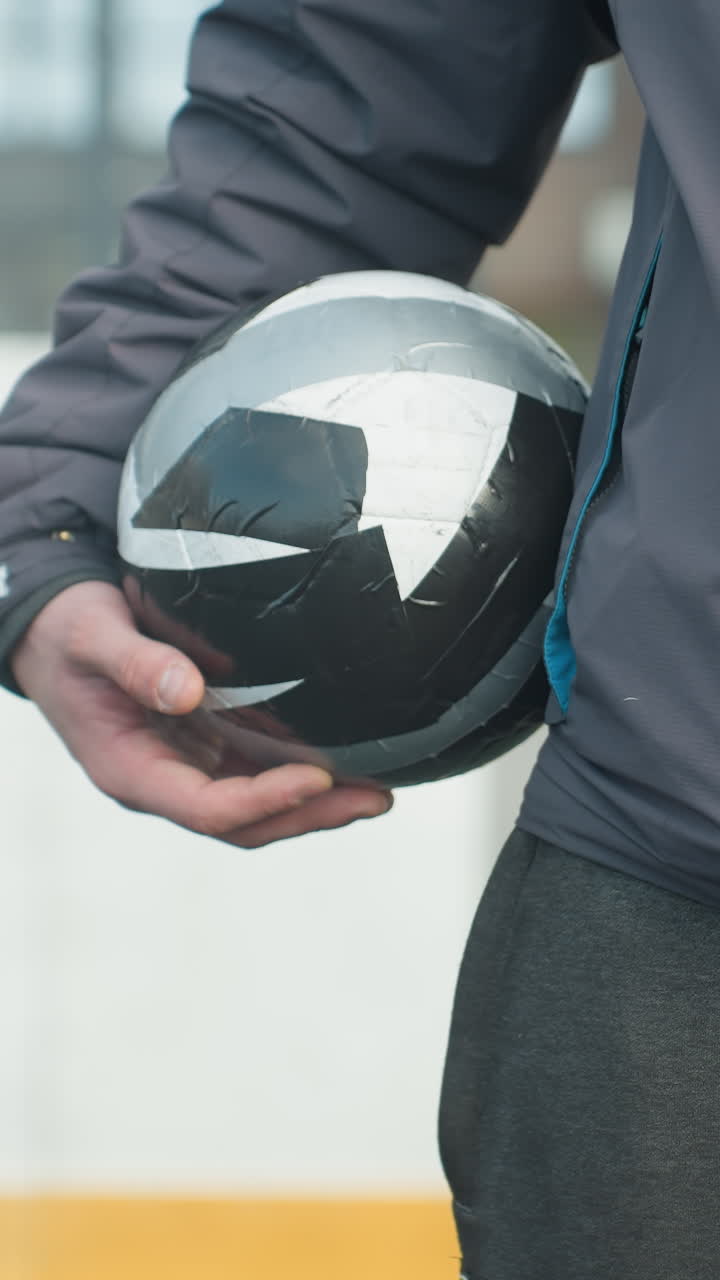Close-up of hand holding black and white soccer ball with focus on player s hand, showing athletic wear and part of body, urban background with buildings and outdoor sports