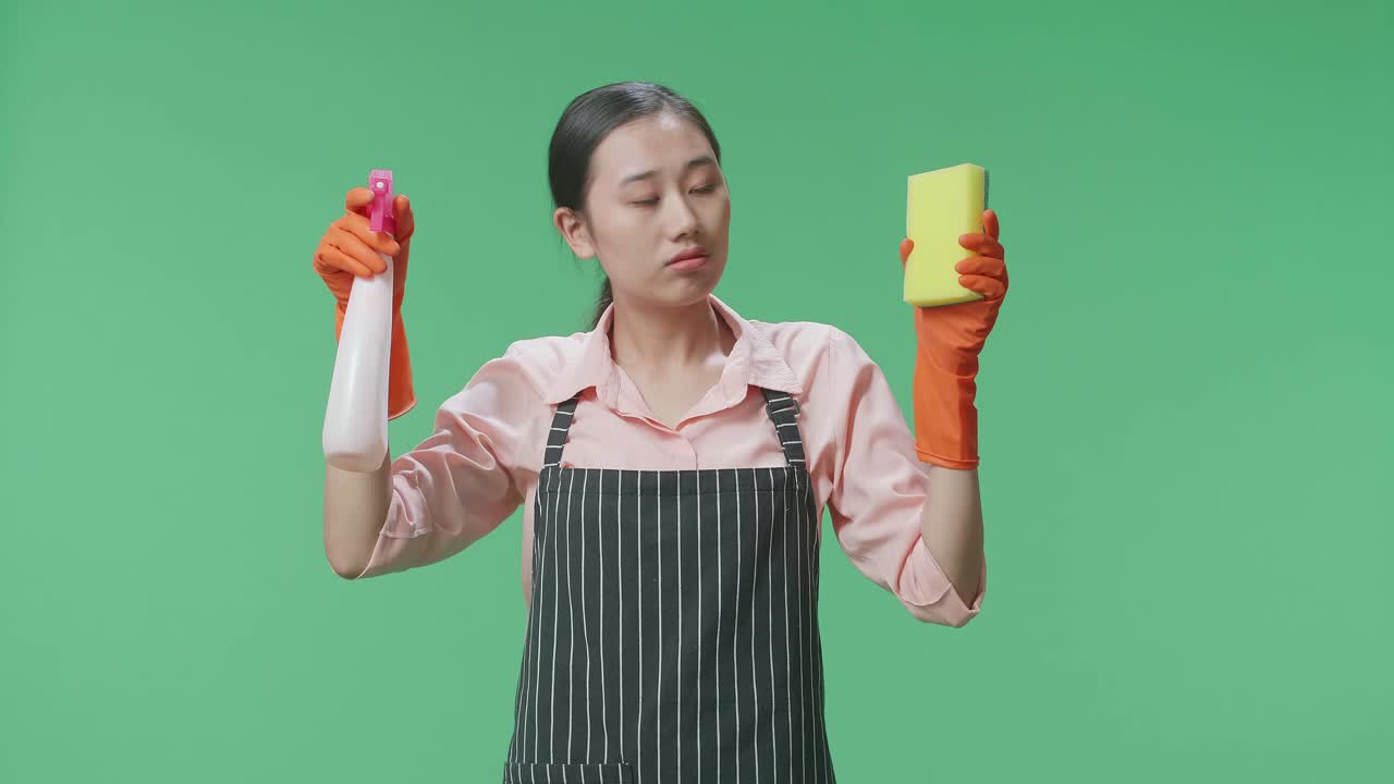 피곤한 아시아 여성 집주인이 스프레이와 주방 스폰지를 손에 들고 초록색 스크린 스튜디오에서 머리를 흔들고 있습니다.