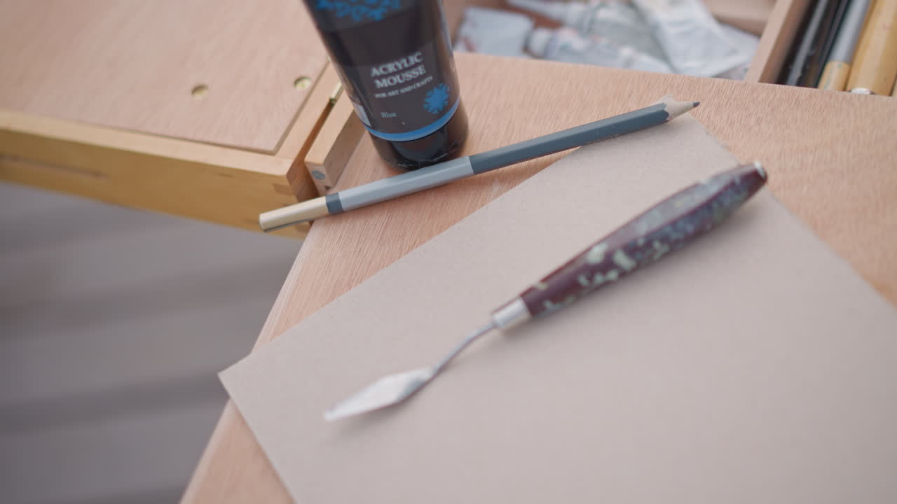 palette knife rests on paper next to paint tube and pencil, closeup on textured surface, studio prep for impasto techniques, metallic blade with paint residue, soft shallow depth, concentrated mood