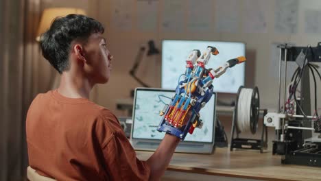 back view of asian boy with 3d printing checking a cyborg hand while designing it on a laptop at home