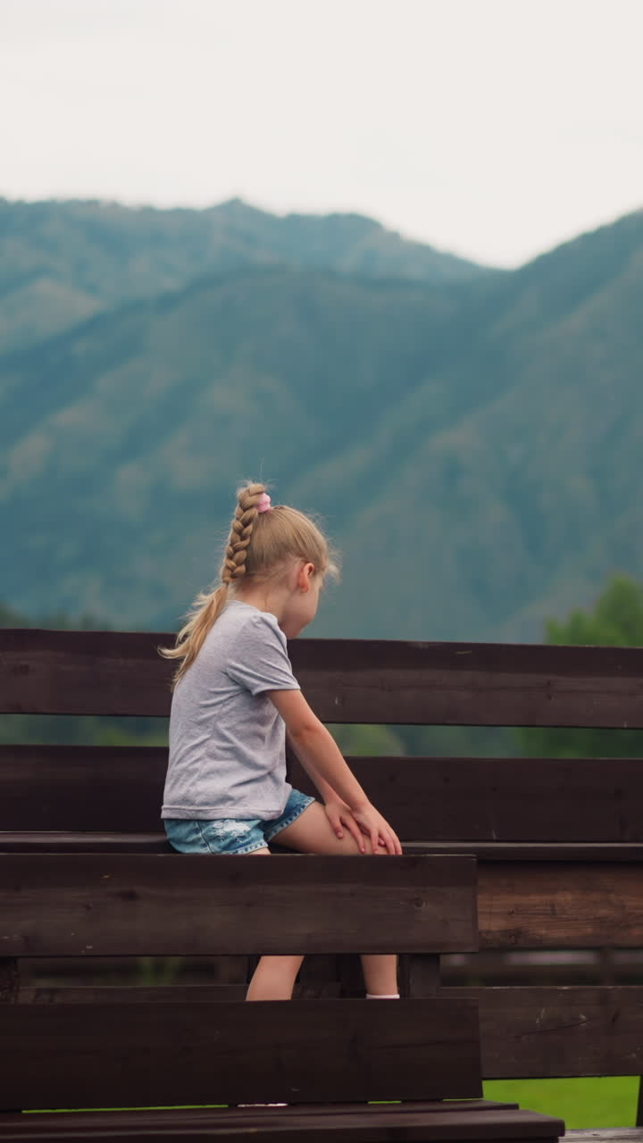 Cute little boy with wild flowers bouquet looks at distant mountains while girl sister sits on wet wooden bench at resort hotel on nasty day