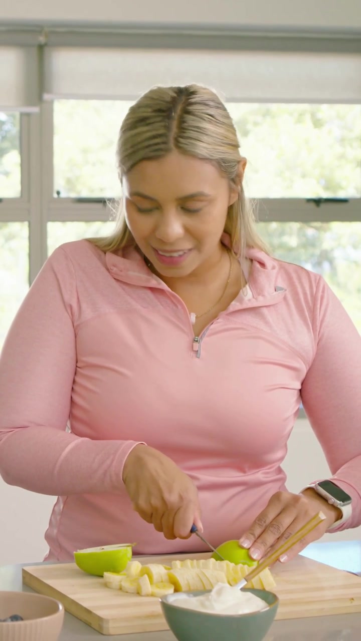 Video verticale: Donna con una felpa rosa con cerniera che prende un coltello e taglia una mela a fette su un tagliere per una ciotola di yogurt.