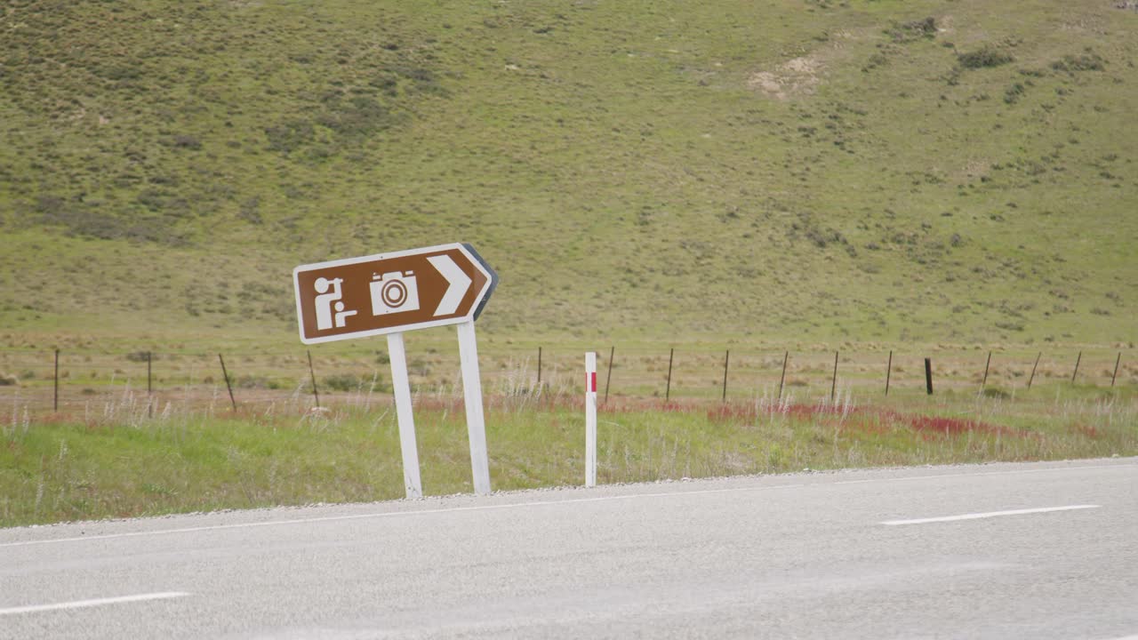 A static shot of a crooked, brown sign next to a road in New Zealand pointing to a lookout spot for taking pictures. Cars drive past the camera