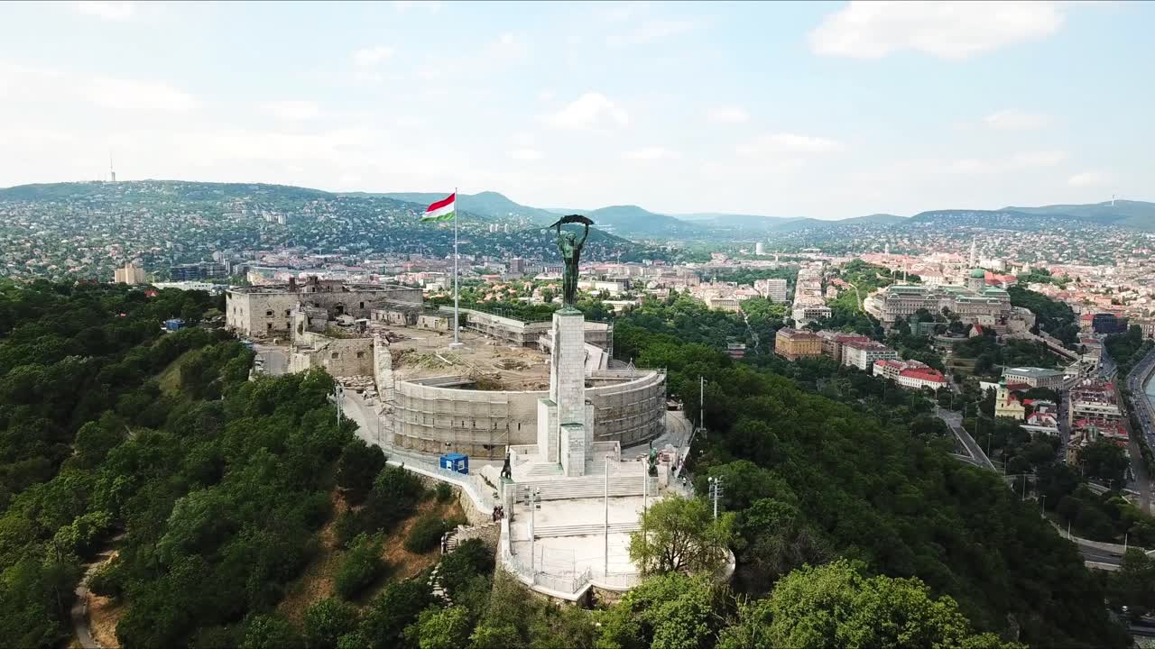 Aerial view of the liberty statue and the citadel on Gellért hill in Budapest, Hungary, showcasing the cityscape and the danube river on a sunny day with the hungarian flag waving, drone orbiting shot
