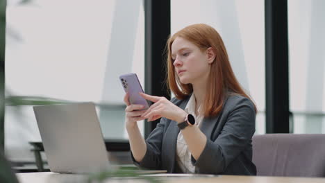 Young-businesswoman-use-smart-phone-with-laptop.-Business-woman-in-a-modern-office-with-a-panoramic-window-sunny-day-laptop-tablet-use-a-mobile-phone-in-a-situation