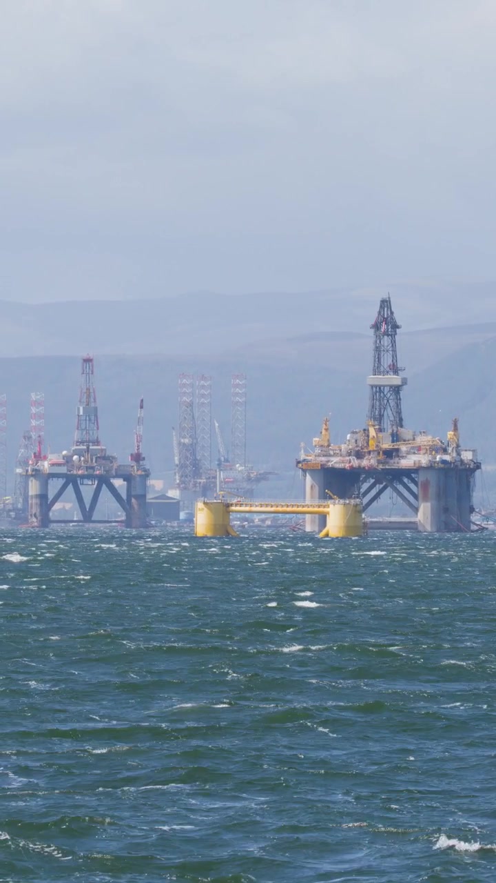 Vidvinkelbillede af en offshore olieboreplatform og en yacht på oprørt hav, dagslys, fjerne skotske højland.