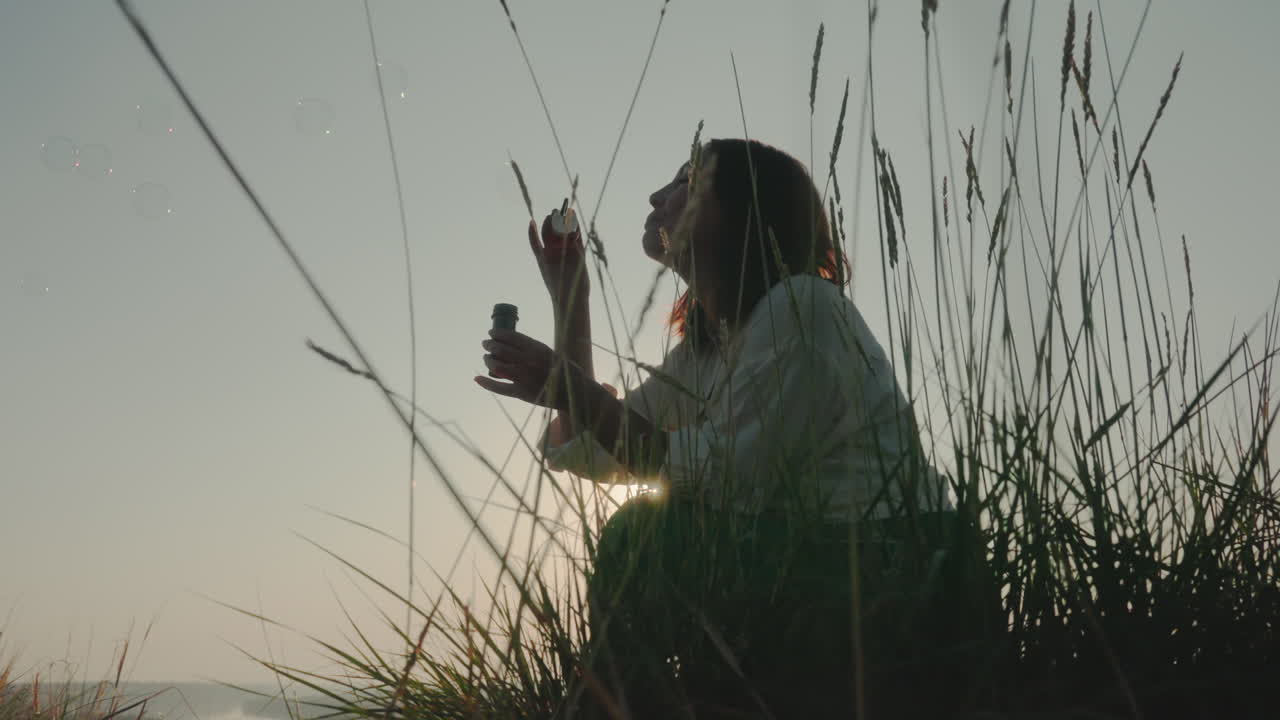 Young crouched among tall wild grass blowing bubbles during soft sunset light, with grain stalks framing silhouette and distant horizon visible in calm natural landscape