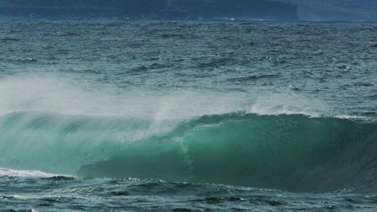 Rolling ocean swell crests and foams at edge as offshore winds lift mist and curve wave face