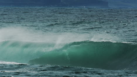 rolling ocean swell crests and foams at edge as offshore winds lift mist and curve wave face