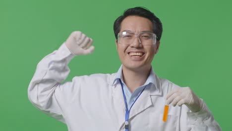 close up of asian man scientist with orange liquid in the test tube smiling and flexing his bicep while standing on the green screen background in the laboratory