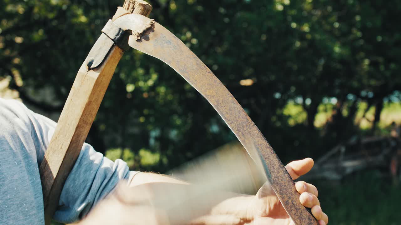 Man sharpening a scythe blade by hand in a rural field. Traditional farming activity captured in close-up with natural light and scenic countryside, green trees background.