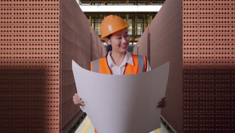 asian female engineer with safety helmet looking at blueprint in her hands and looking around while standing with red brick packed in stacks are stored