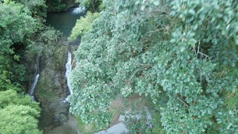 Ascending-drone-view-of-a-woman-in-a-pink-dress-standing-at-the-base-of-a-cascading-waterfall