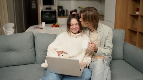 Young-adult-couple-talking-using-laptop-computer-at-home-sitting-on-couch,-watching-video