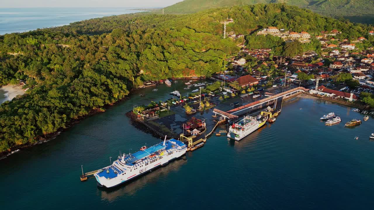 Ferries waiting at Padang Bai Port in Bali as a drone view captures the calm sea, nearby boats, and green hills surrounding this active coastal gateway between islands.