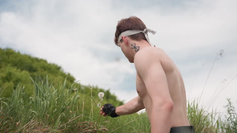 shirtless soldier with visible neck tattoo and white headband climbs grassy hillside reaching to pluck wildflowers, surrounded by cloudy sky and green foliage, captured in natural outdoor lighting