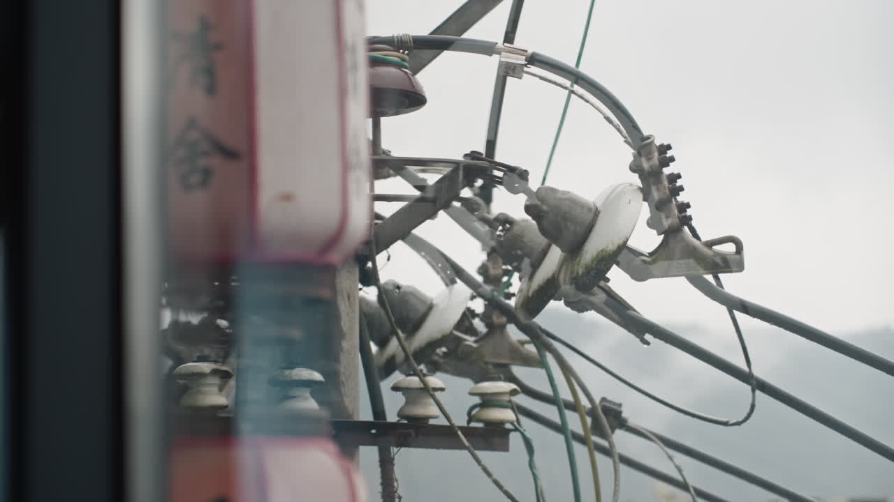 Overcast urban power pole tangled cables, seen through window frame, close shot of transformer and insulators, raindulled sky and distant hills, muted color palette, industrial mood with detailed