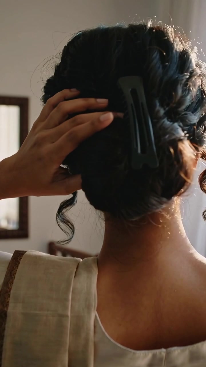 Vertical video: Lifting right hand, woman in saree pallu fixing bun by window with large black clip