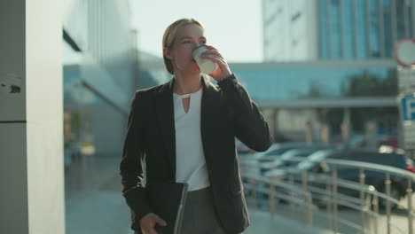 young woman in formal outfit walking outdoors holding tablet in one hand and takeaway coffee in other hand with warm smile on face, surrounded by parked cars and modern glass buildings