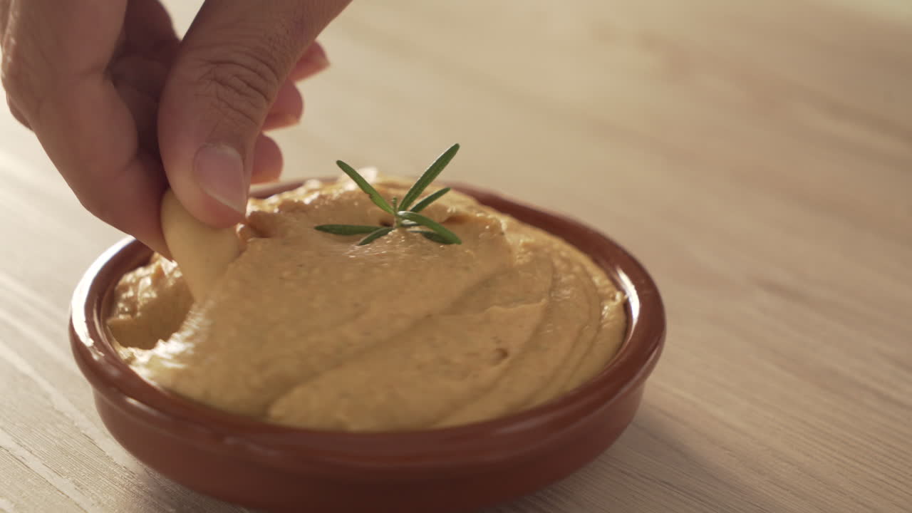 Dipping a Cracker into Hummus with Rosemary Garnish