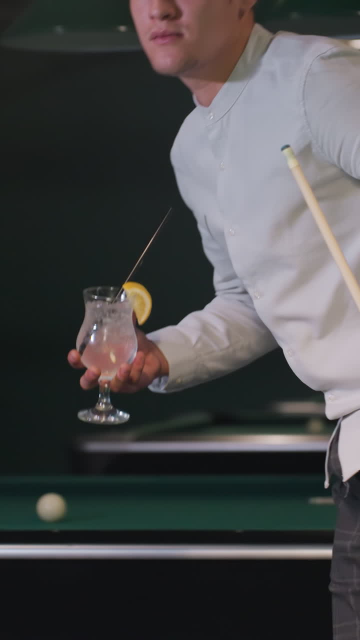 Primo piano di un uomo con camicia bianca e pantaloni grigi che tiene in mano una stecca da biliardo e un bicchiere di limonata mentre passeggia in una sala da biliardo. Le luci dall'alto creano un'atmosfera rilassata ed elegante sopra i tavoli da biliardo verdi.