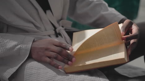 man in white robe reading book, turning pages while sitting in peaceful bedroom. sunlight gently illuminates his hands, creating a serene and focused atmosphere for quiet reflection and reading