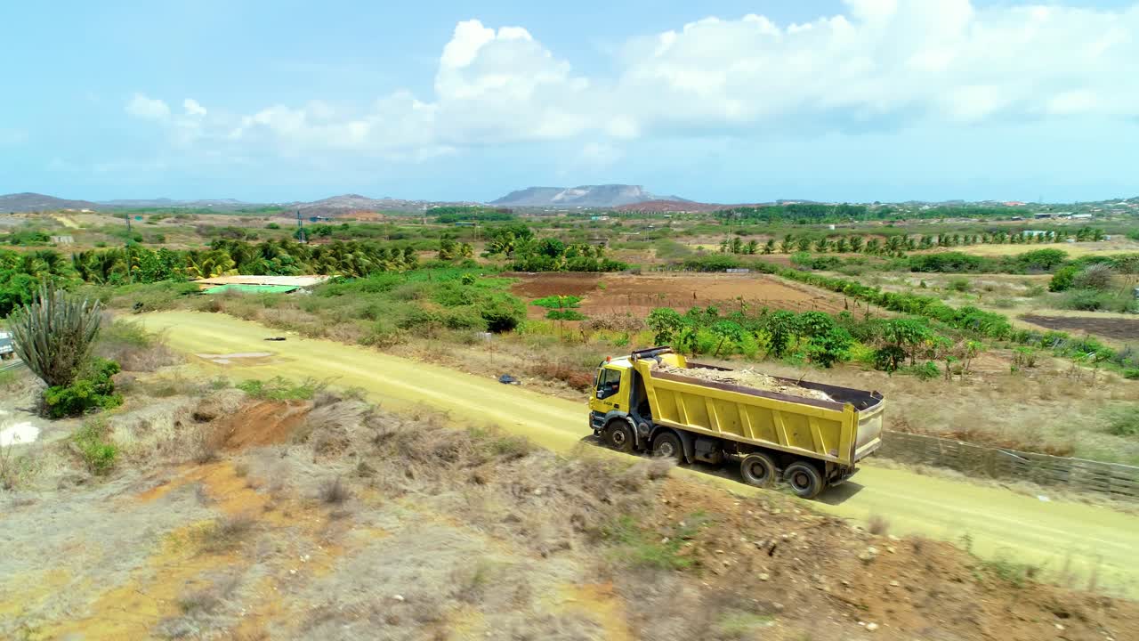 4k aerial cinematic follow orbit shot of yellow dump truck filled with dirt and rubbish, driving through rural farmland on a dirt road during a beautiful sunny day in the Caribbean, Curacao