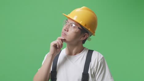 close up de hombre asiático trabajador con gafas de protección y casco de seguridad pensando en algo y mirando a su alrededor mientras está de pie en la pantalla verde estudio de fondo