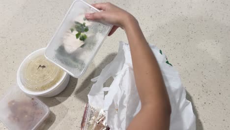 hands unpacking takeout food containers from a plastic bag on a kitchen counter under bright lighting