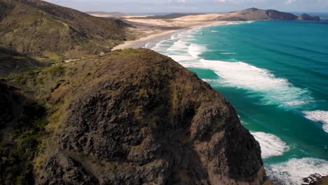 hombre de pie en una colina admirando las olas del océano rodando en la playa de arena de te werahi, parque nacional en northland, nueva zelanda