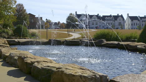 public park pond area with several fountains spraying streams of clear water with homes and buildings in the background, on a sunny day