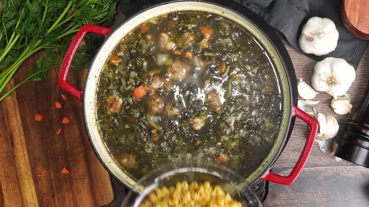 Cooking Italian Wedding Soup while adding in the ditalini pasta into the red stock pot. Mixing in small pasta into the pot while it is simmering. Using a wooden spoon to stir the hot soup.