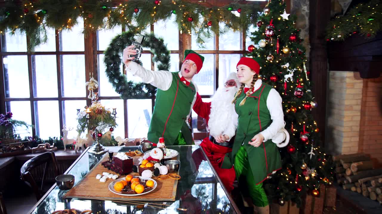 Funny elves and Santa in costumes in Santa's residence.Two elves and santa Claus are posing to retro camera at Christmas