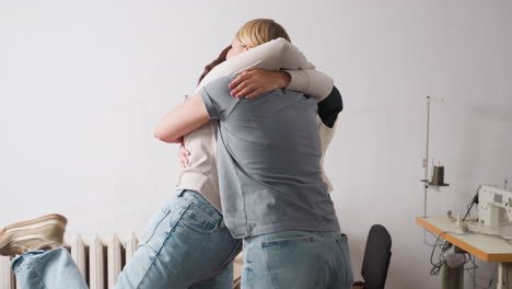 Creative women in sewing studio leap into joyous hug, arms wide, celebrating project success beside industrial machines, denim jeans swishing, cheerful energy filling minimalist workspace