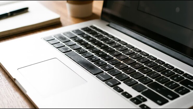 Nahaufnahme einer Laptop-Tastatur auf einem Holzschreibtisch mit Notizbuch, Stift und Kaffeetasse bei sanftem natürlichem Licht.