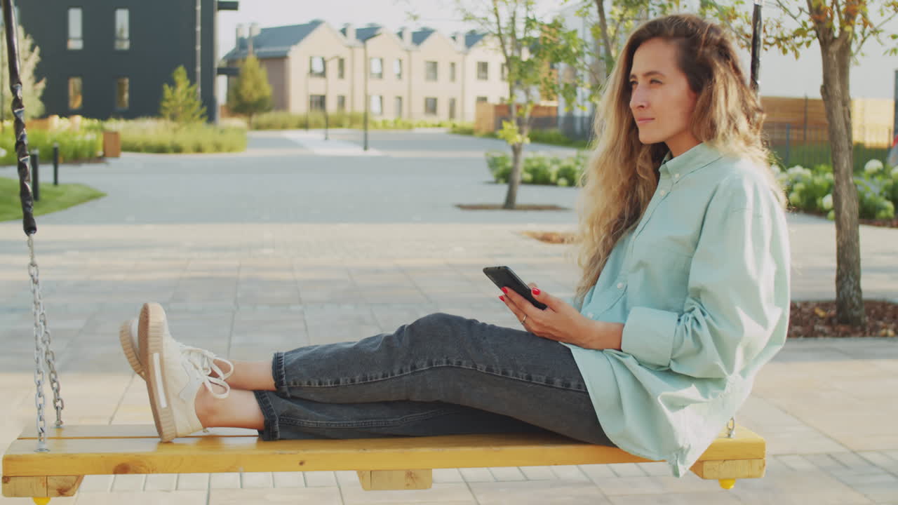 mujer sentada en un columpio al aire libre y usando un teléfono inteligente