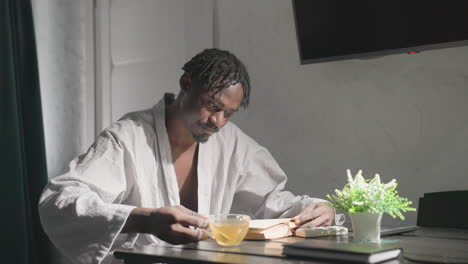 man in white robe sitting at table in cozy, modern living space sipping tea while reading book. bright sunlight and soft lighting create calm and relaxed atmosphere perfect for unwinding