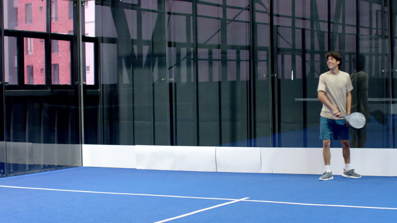 Young man playing padel tennis on indoor court, smiling and holding racket