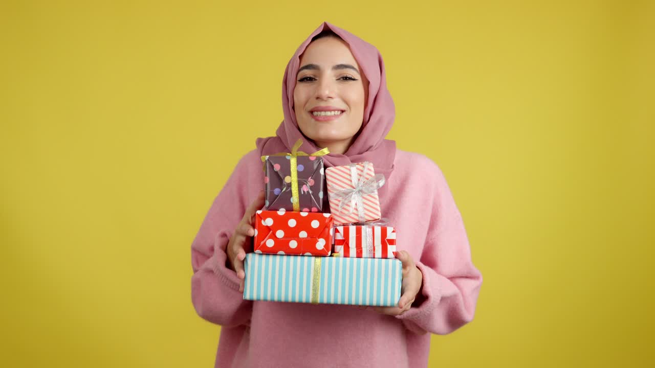 Happy Woman in Hijab Holding a Stack of Gifts on Yellow Background