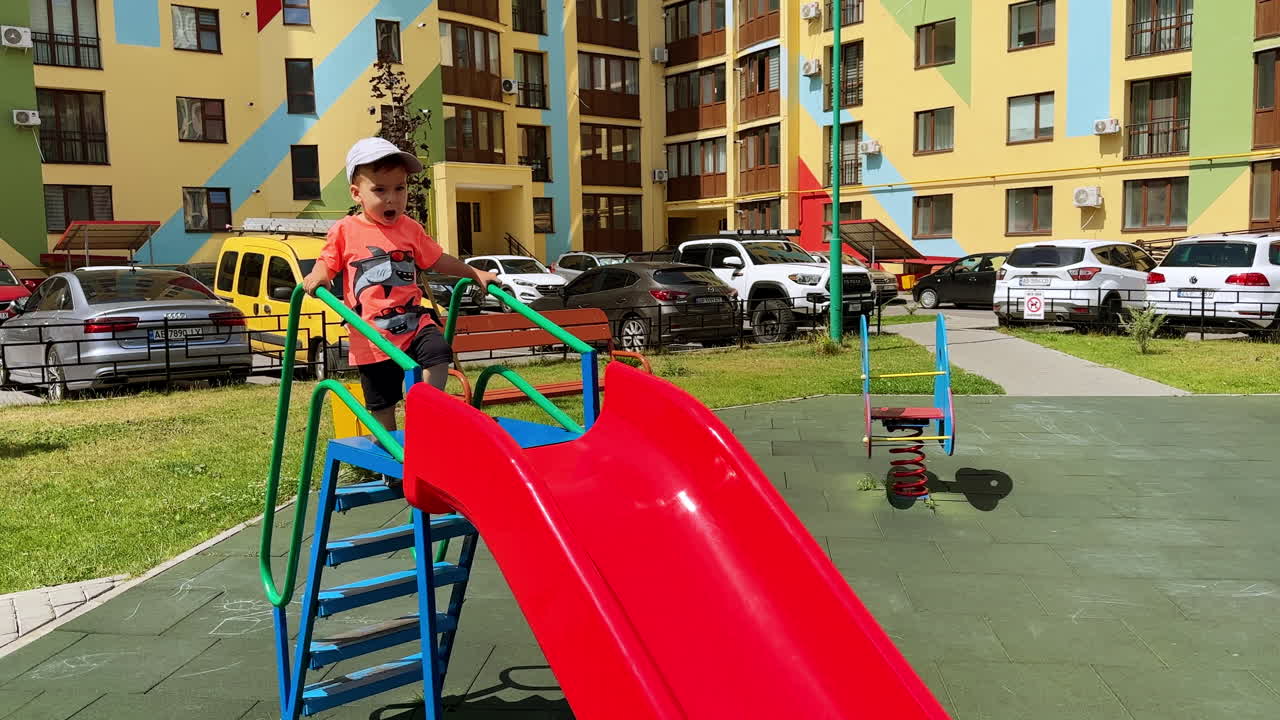 Talkative baby boy is on the slide outdoors. Toddler in cap slides down and claps hands cheerfully.