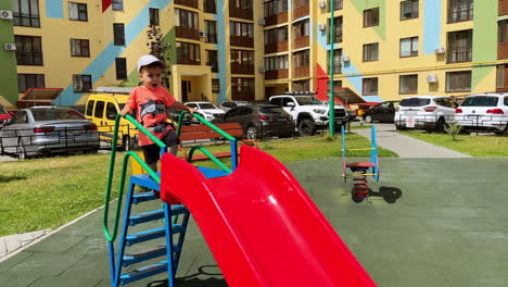 talkative baby boy is on the slide outdoors. toddler in cap slides down and claps hands cheerfully.