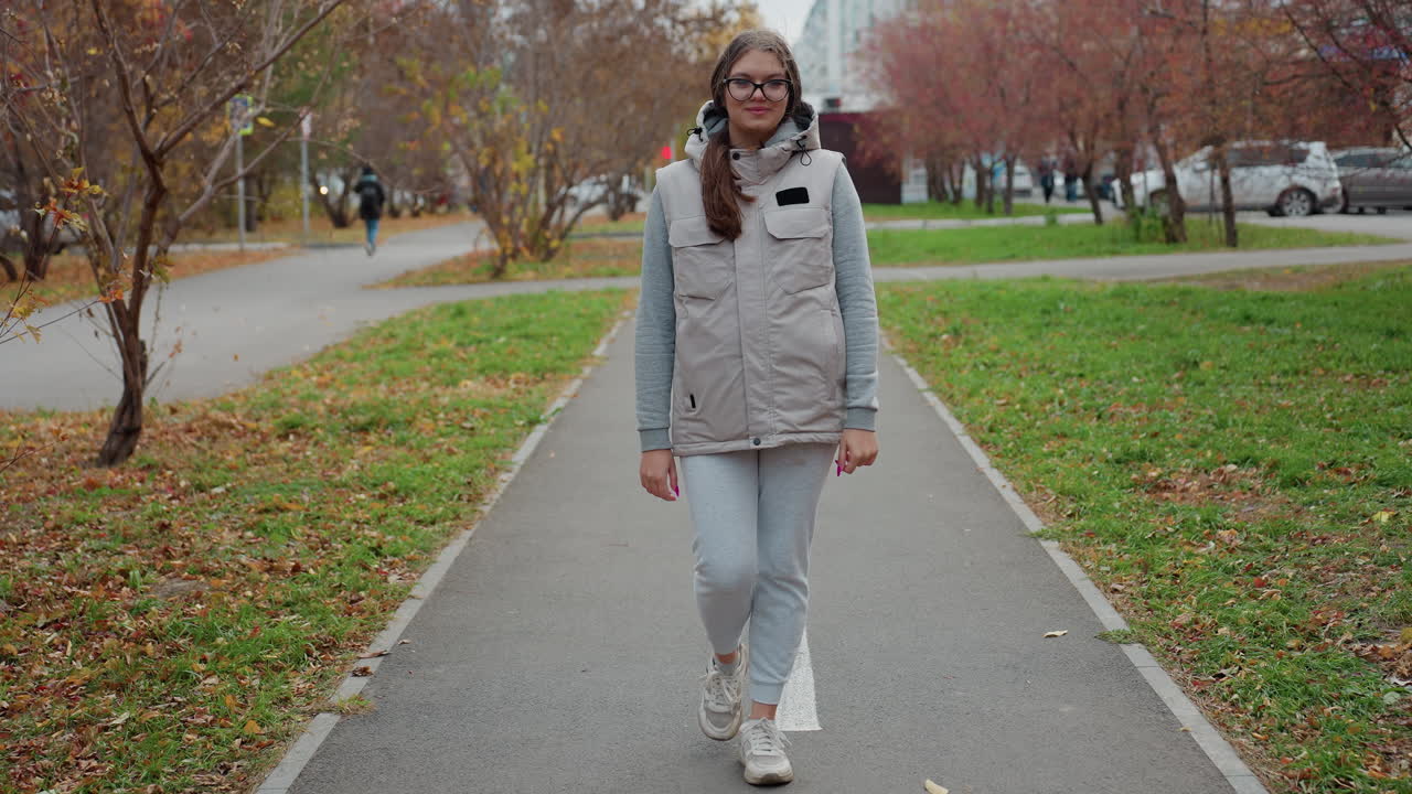 Fashion lady dressed in joggers walking majestically in busy urban park lined with autumn trees and fallen leaves as people move in background and cars drive past residential buildings nearby