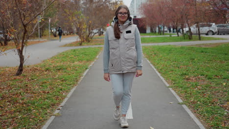 fashion lady dressed in joggers walking majestically in busy urban park lined with autumn trees and fallen leaves as people move in background and cars drive past residential buildings nearby