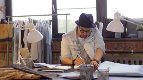 joven diseñador de moda masculino sentado en el escritorio, usando una computadora portátil y dibujando un boceto de un vestido nuevo en un moderno estudio de costura 1