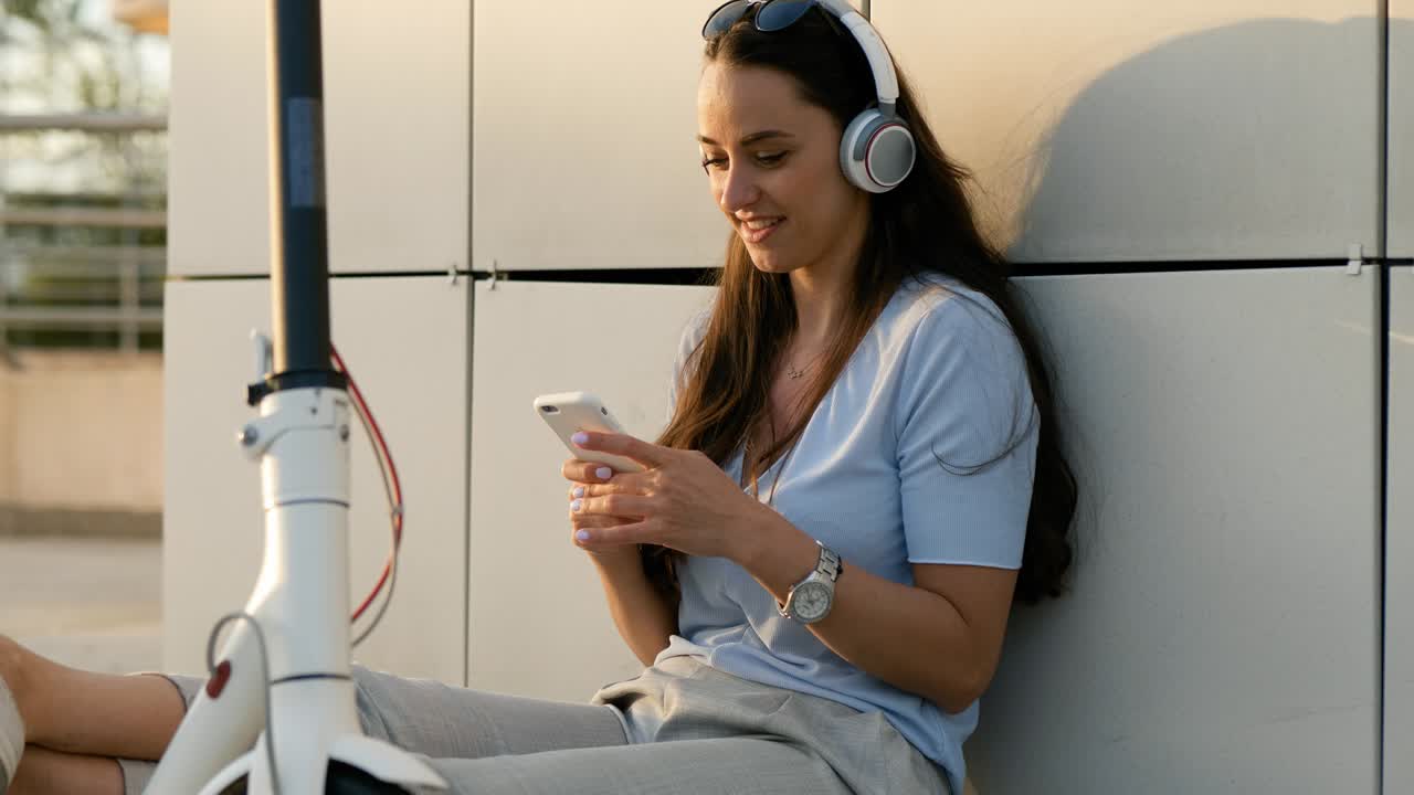 Young beautiful girl listening to music in her smartphone, sings  while sitting next to an electric kick scooter