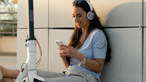 young beautiful girl listening to music in her smartphone, sings  while sitting next to an electric kick scooter