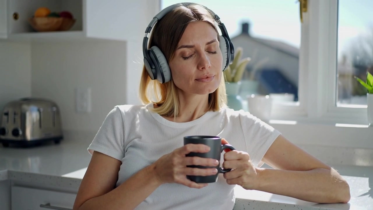 Donna seduta che indossa una cuffia, tiene una tazza al bancone della cucina mentre la luce del sole filtra dalla finestra.
