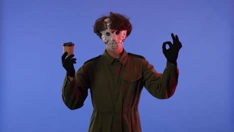 man in white mask with cup of coffee shows thumbs up gesture on blue background with red neon light