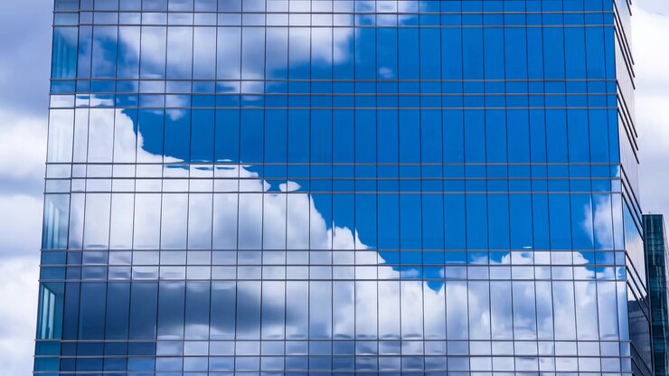 Cielo blu e nuvole bianche a strati scivolano attraverso i pannelli riflettenti delle finestre sull'esterno di un moderno grattacielo, mentre la griglia fissa crea linee nitide e forme speculari mutevoli.